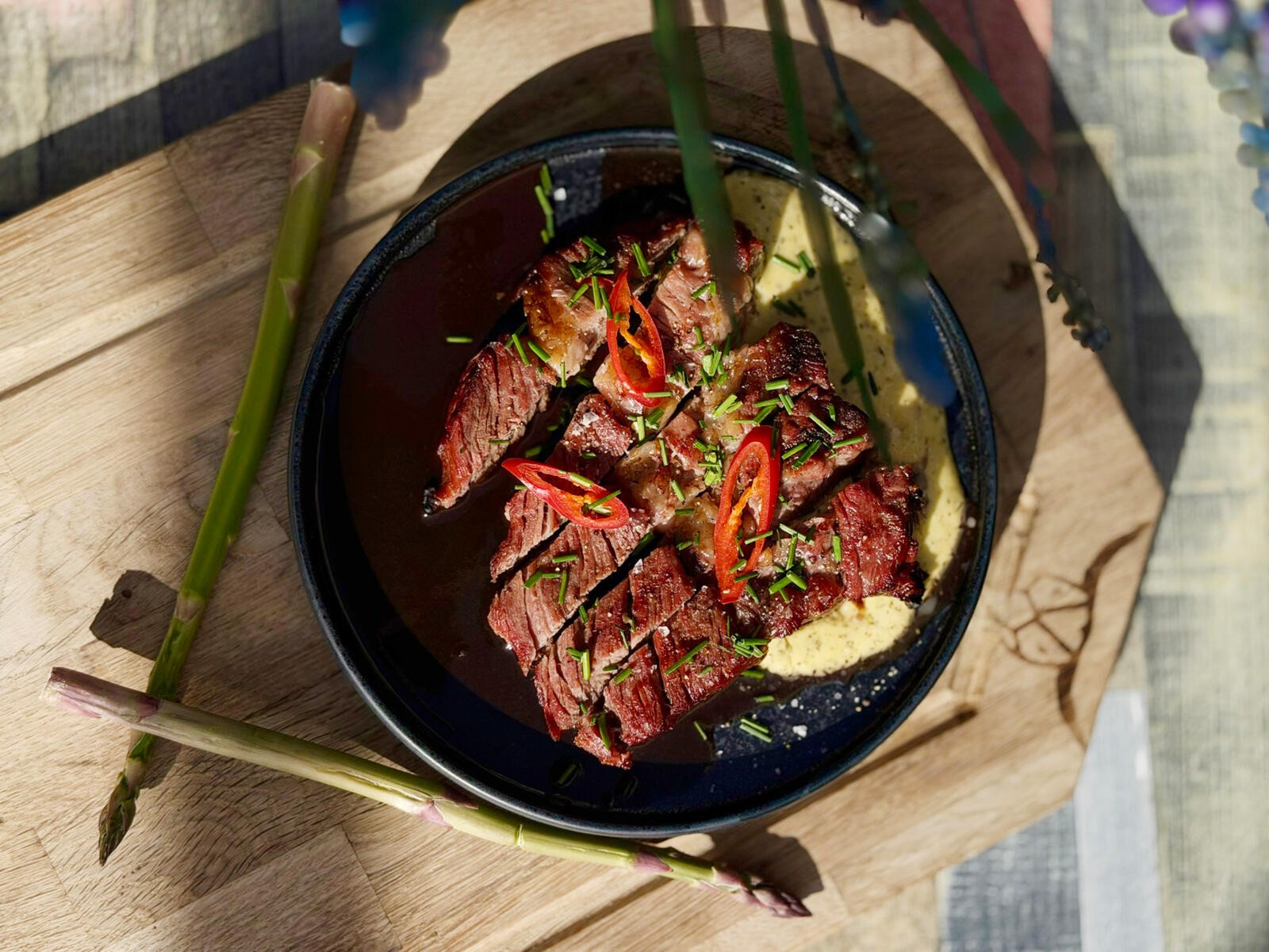 Dark bowl with sliced grilled meat garnished with chili and chives, served on wooden board with asparagus shoots.