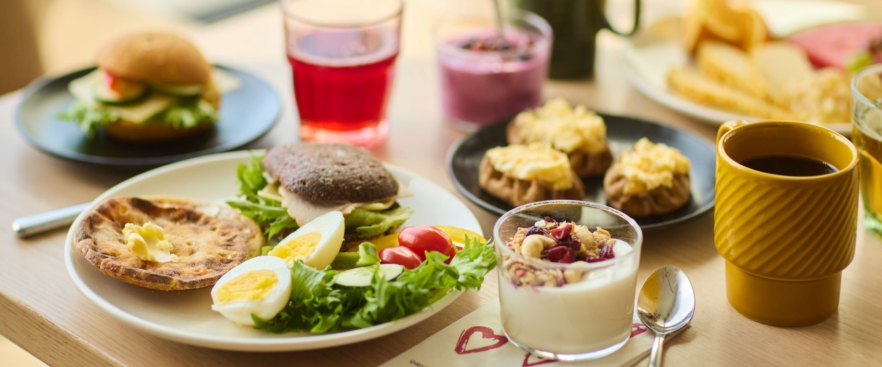 Monipuolinen aamiainen pöydällä: salaattia, munia, burgereita, leipää, dumplingeja, smoothie, jogurtti ja kahvia.