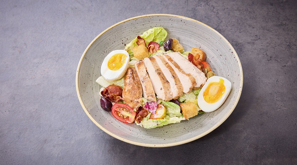 Annos salaattia, jossa kanaa, kananmunaa, krutongeja, pekonia ja tomaattia vaaleanharmaalla keraamisella lautasella.