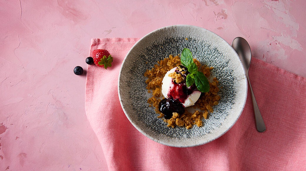 Dessert in a patterned bowl: vanilla ice cream, berry sauce, crumble and mint. A spoon, strawberry and blueberries lie on a pink surface.