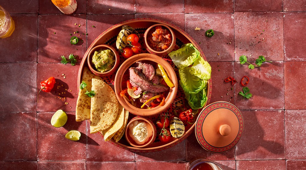 Terrakotatald grillitud liha ja paprikaga, tortillad, guacamole, salsa, kaste ja salatilehed punasel plaatpinnal.