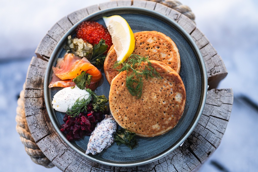 Bliniannos sinisellä lautasella puisella alustalla. Tarjoillaan lohen, mädin, smetanan, sitruunan ja mausteyrttien kera.