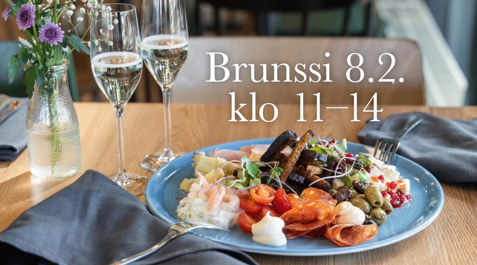 Sinisellä lautasella antipastilajitelma sisältäen katkarapuja, salamia, oliiveja ja muita herkkuja. Vieressä kaksi lasillista kuohuviiniä ja kukkamaljakko.