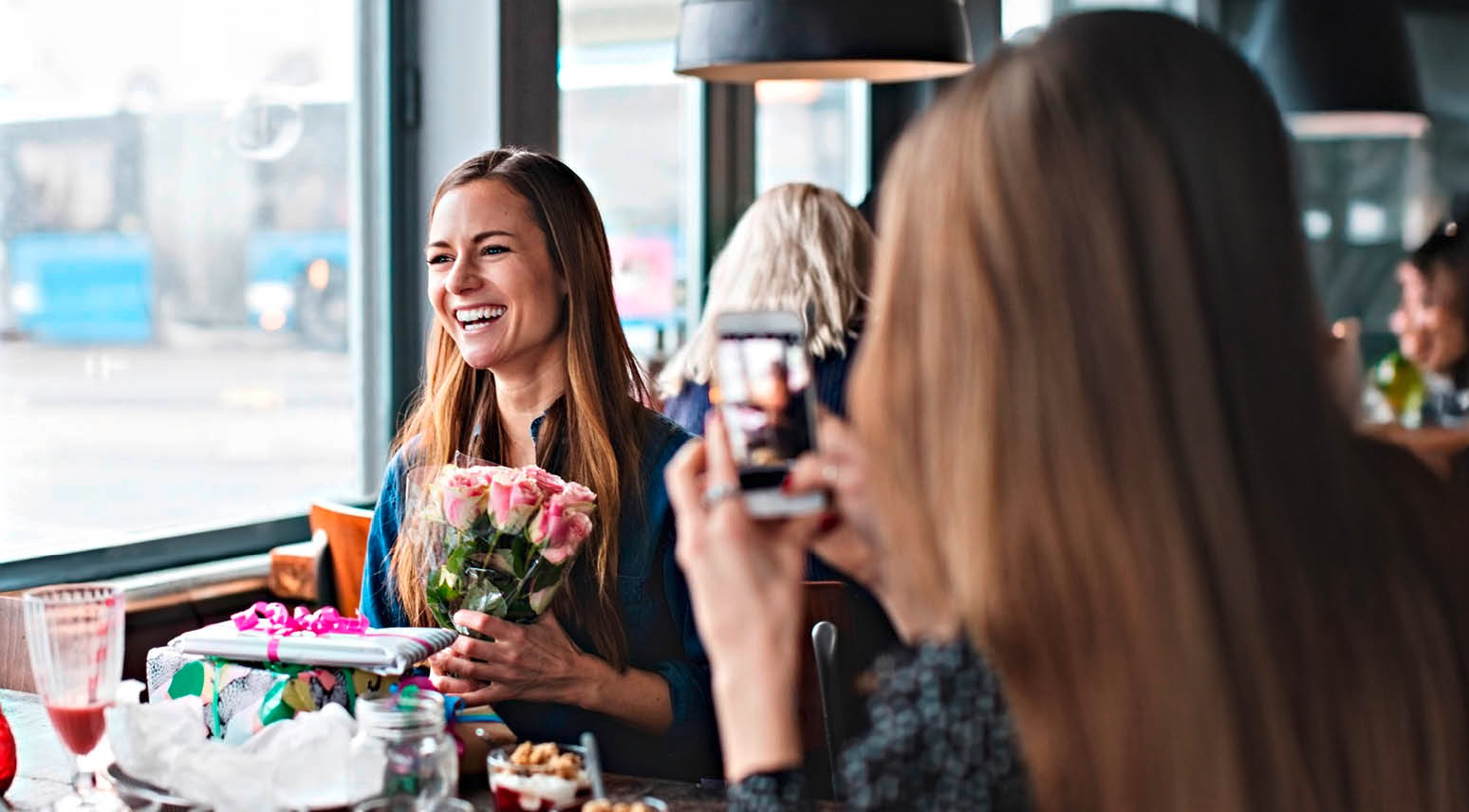 Person håller rosa rosor och presentpaket på ett kafé medan någon tar ett foto. Drycker och efterrätter på bordet.