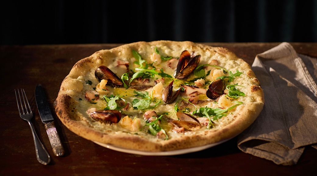 Skaldjurspizza med musslor, räkor och rucola på ett träfat. Bredvid ligger gaffel, kniv och servett.