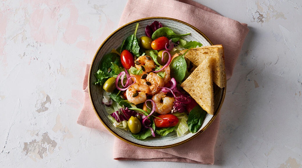 Prawn salad in a bowl with leafy greens, cherry tomatoes, olives, red onion and pumpkin seeds. Toasted bread triangles on the side.