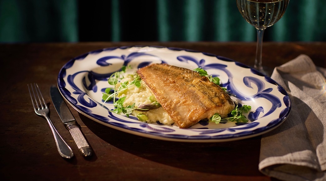 Praetud kala risoto peal sinivalge dekoratiivse taldrikul, kõrval veinipokaal ja söögiriistad.