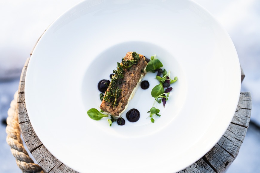 Tyylikkäästi asetettu ruoka-annos valkoisella lautasella: kalafilee yrttikoristuksella, mustia kastikepisteitä ja tuoreita yrttejä ympärillä, puisella alustalla.