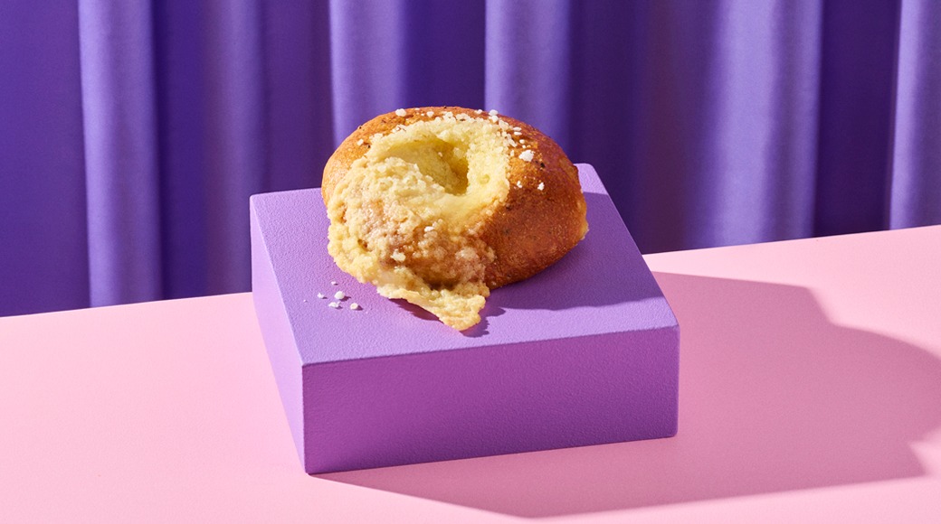 Pulla, josta valuu vaaleaa täytettä, violetilla alustalla vaaleanpunaisella pöydällä. Taustalla violetti verho.