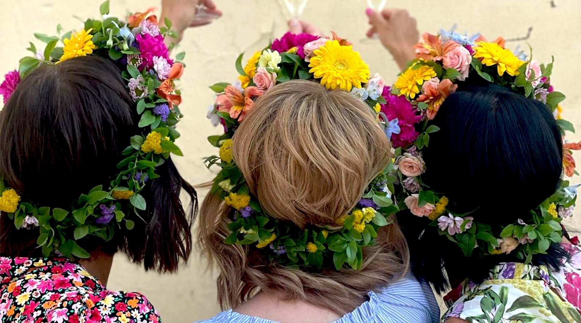 Tre personer med blomsterkransar på huvudet, fotograferade bakifrån. Kransarna har färgglada blommor som gerbera och rosor.