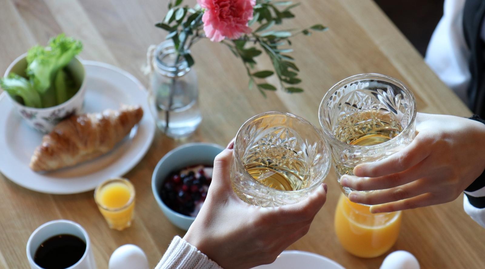 Hands toasting crystal glasses over breakfast table with croissant, berries, orange juice and pink flower in vase.
