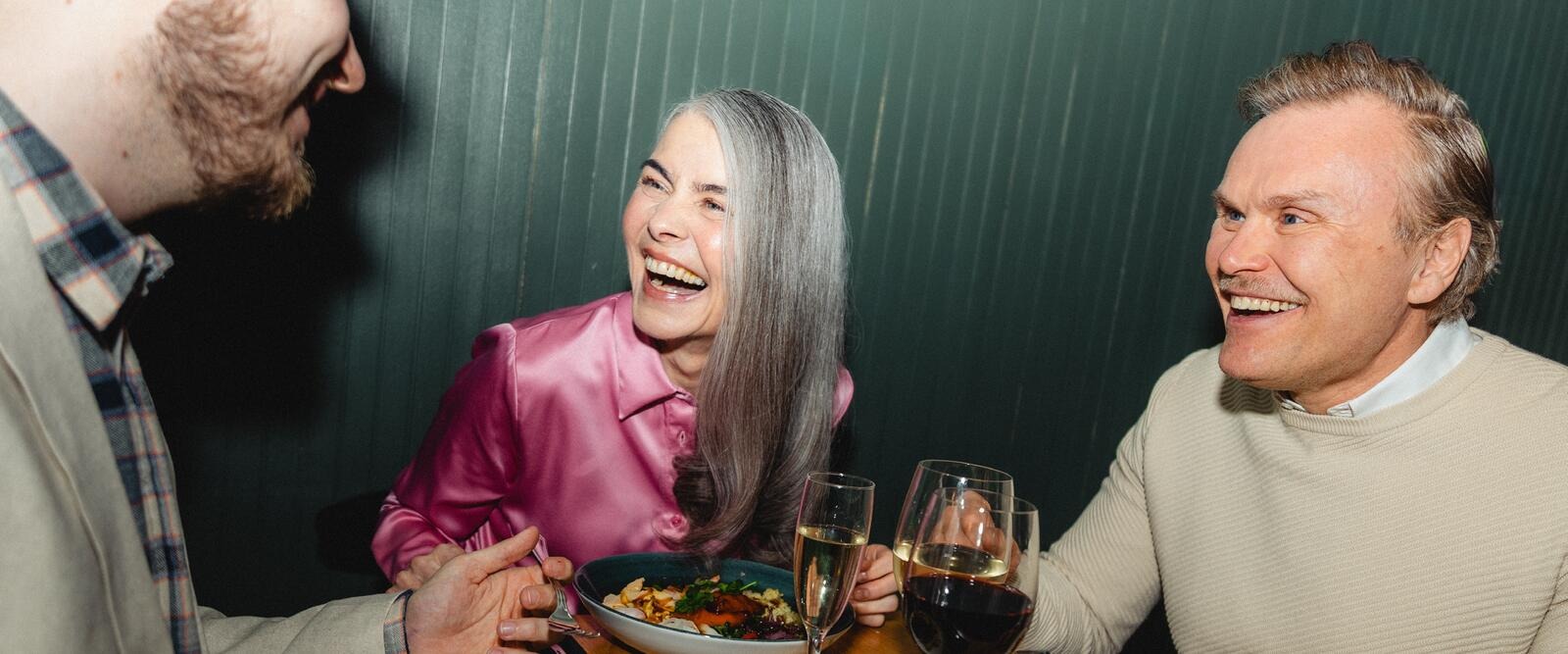 Three people sitting at a cushioned bench in a restaurant, laughing and conversing. Food and drinks on the table.