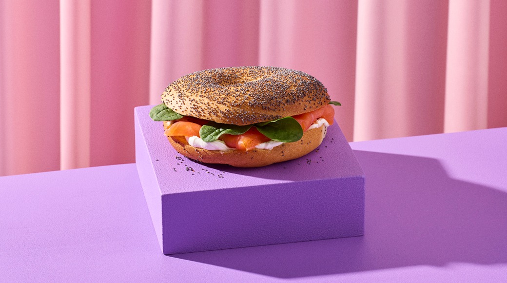 Unikkopäällysteinen bagel, jonka välissä on savulohta, tuorejuustoa ja pinaatinlehtiä. Bagel on violetin korokkeen päällä vaaleanpunaista verhoa vasten.