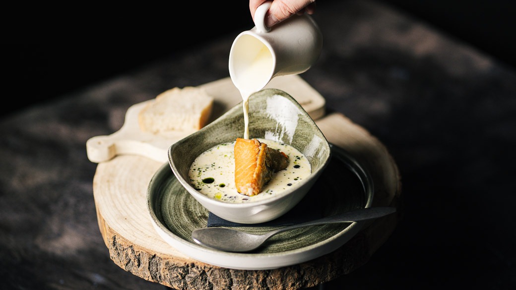 Käsi kaataa kermaa kulhoon, jossa on keitto ja rapea leipäpala. Annos on tarjoiltu puisella leikkuulaudalla.