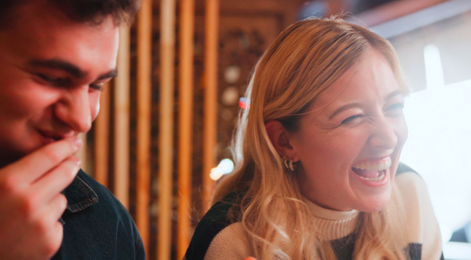Kaksi henkilöä nauravat yhdessä lämpimässä sisätilassa, jossa on puisia seinäelementtejä taustalla.