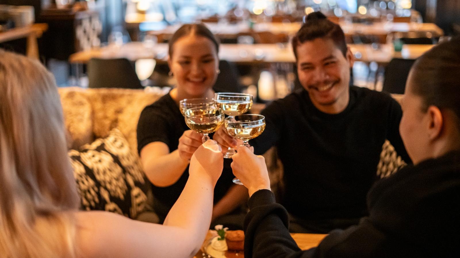 Four people toasting with champagne glasses in a restaurant. A cozy, dimly lit dining area is visible in the background.