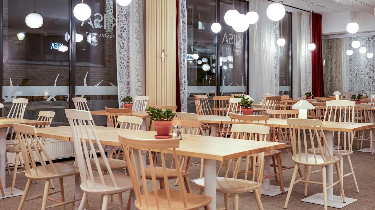 Vaalean puun ja valkoisen sävyisiä neljän ja kuuden hengen pöytäryhmiä suurten ikkunoiden äärellä.