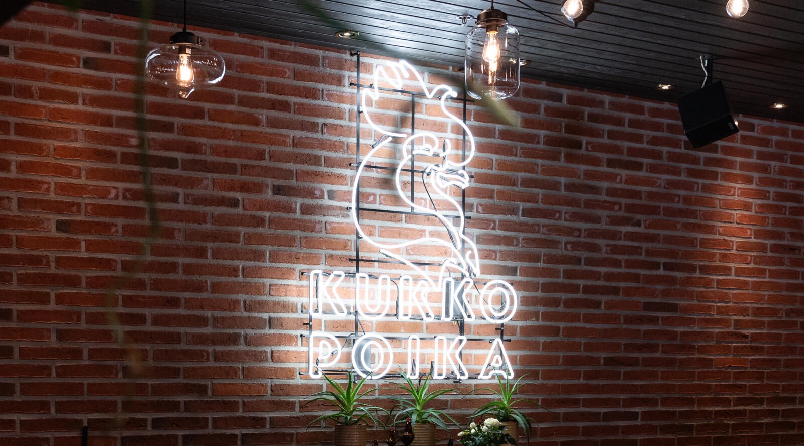 Tiiliseinällä on hohtava valokyltti Kukkopojan logosta.