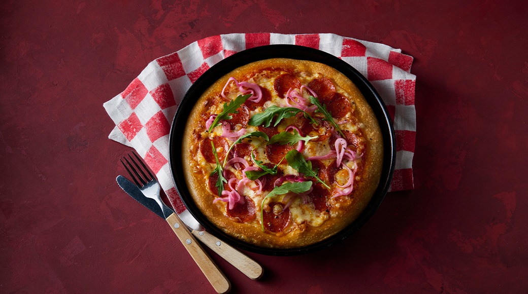 Pepperoni pizza topped with arugula and pink pickled onions in a black pan on a red and white checkered cloth. A fork and knife lie beside it.