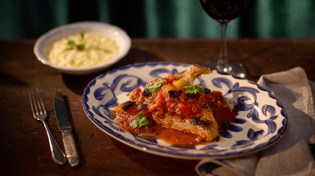 Kana tomati-oliivi kastmes sinisel-valgel taldrikul, riisiga ja punase veini klaasiga puulaual.