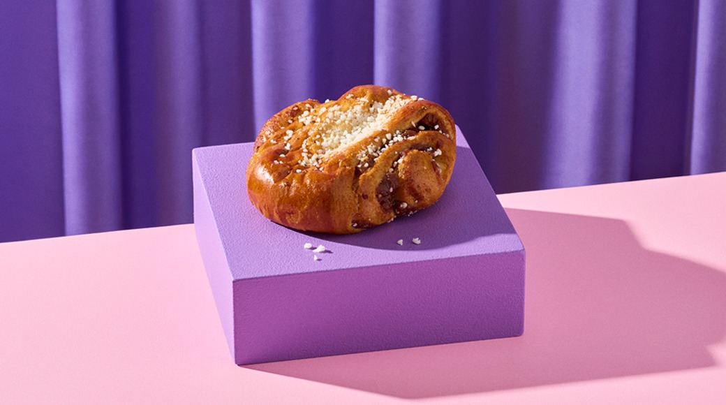 Pulla, jonka päällä on raesokeria, violetilla korokkeella vaaleanpunaisella pöydällä. Taustalla on violetti verho.