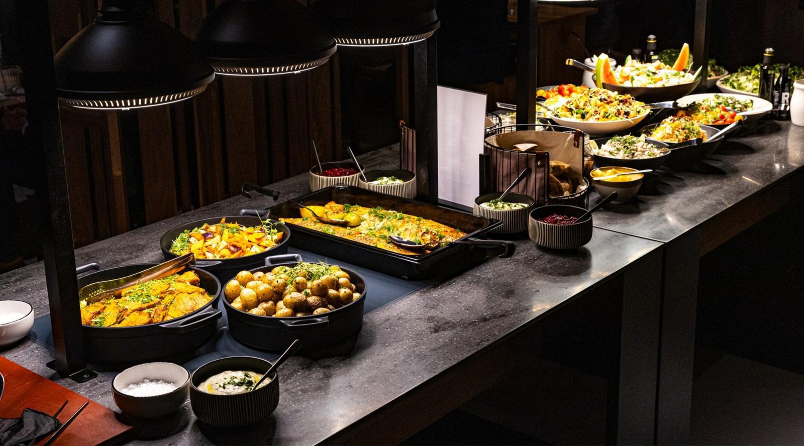 Buffet table displaying a wide variety of dishes in bowls and trays. The table is equipped with warming vessels and three black pendant lamps hang overhead.