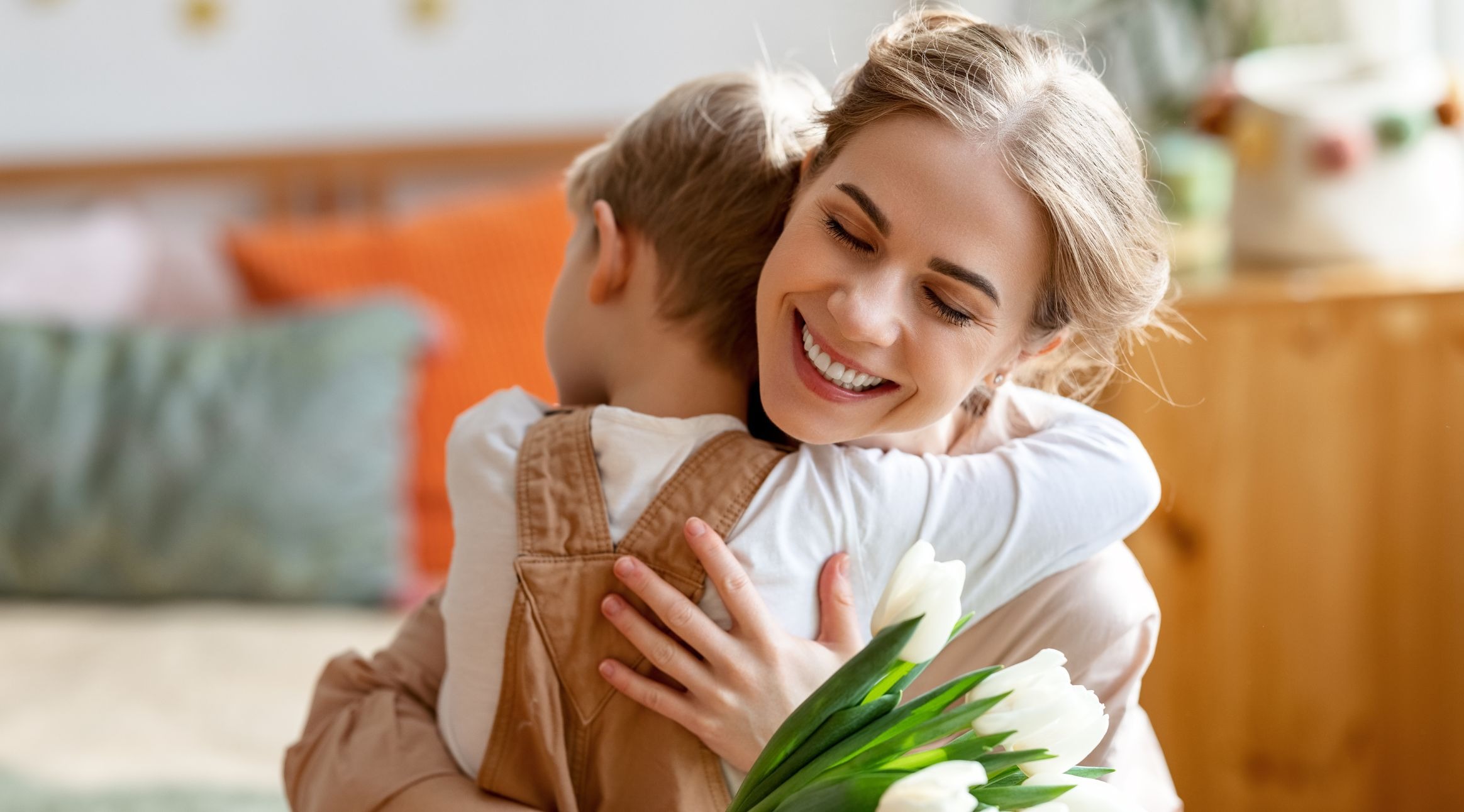 Hymyilevä nainen halaa lasta kodin lämpimässä sisustuksessa, kädessään valkoisia tulppaaneja.