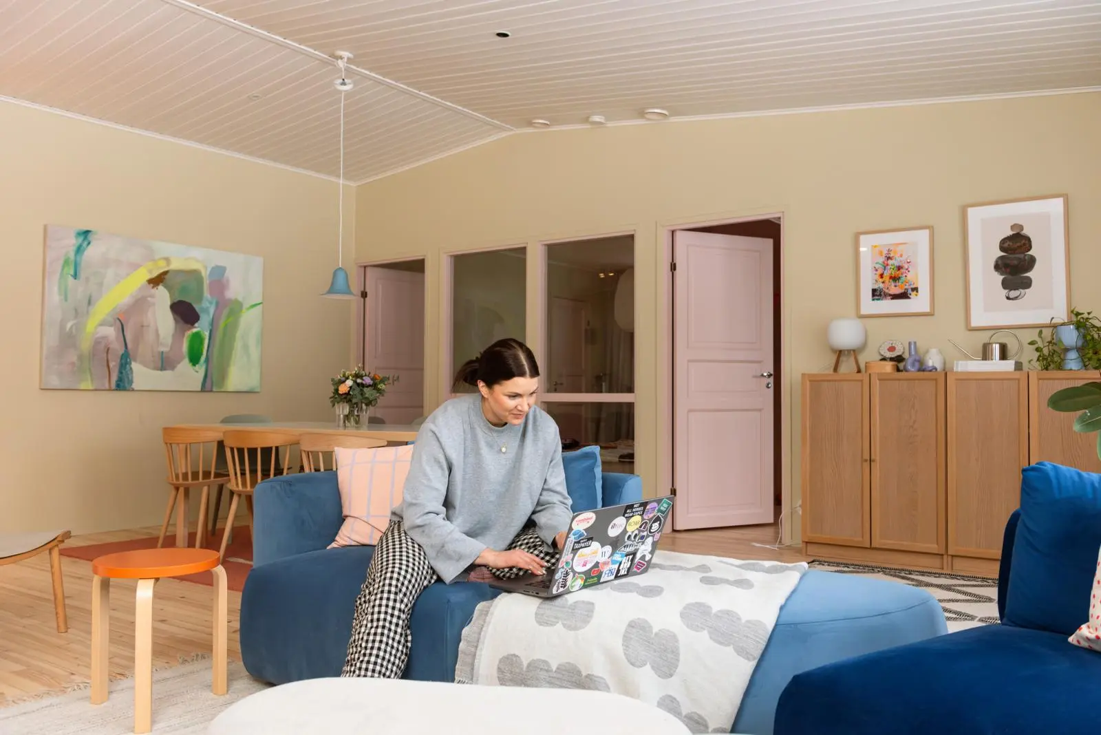 Maru Hautala istuu sinisellä sohvapalalla kannettavan tietokoneen kanssa. Osa taustalla näkyvästä seinästä on lasi-ikkunaa.