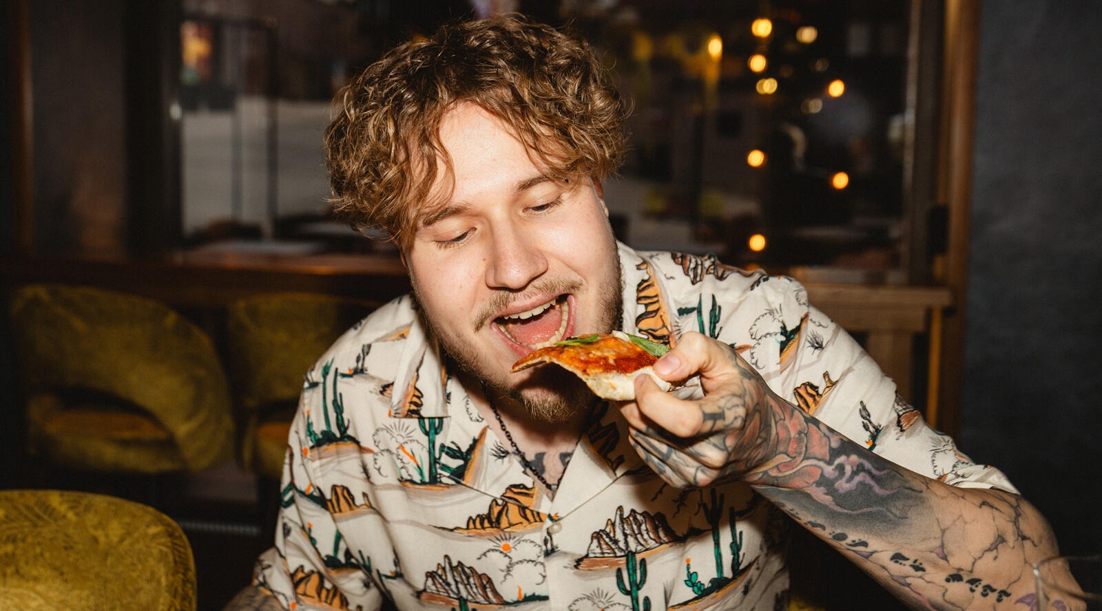 Man eating a slice of pizza on a soft sofa in a restaurant with dim lighting.