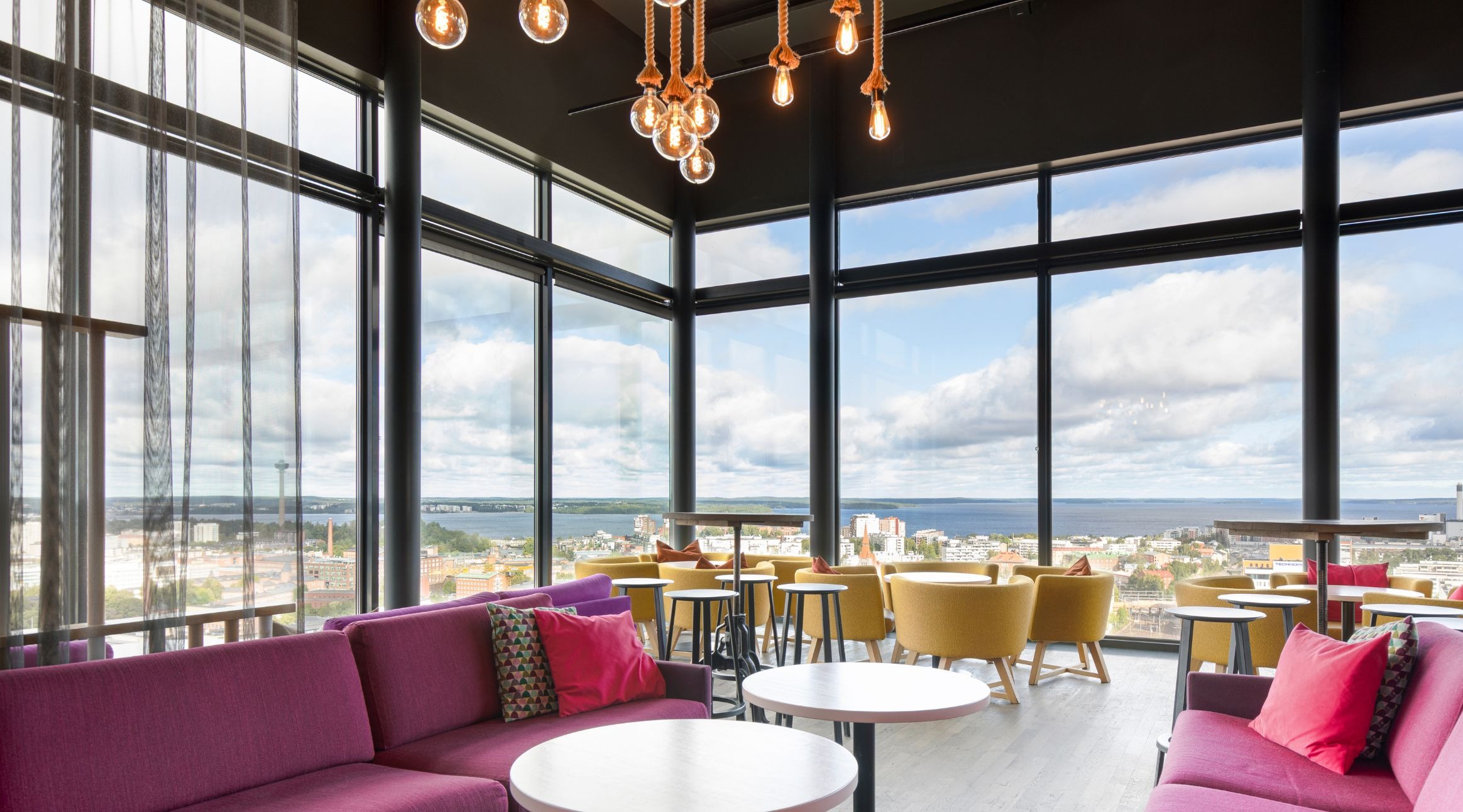 Moderni loungealue, jossa värikkäät sohvat, keltaiset nojatuolit ja panoraamaikkunat tarjoavat näkymän järvelle ja kaupunkiin. Katossa roikkuu koristeelliset hehkulamput.