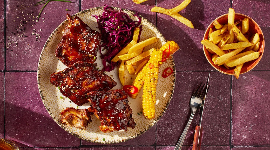Lautasella glaseerattuja grillattuja ribsejä seesaminsiemenillä, punakaalisalaattia, ranskalaisia ja maissintähkiä. Vieressä kulho ranskalaisia sekä haarukka ja veitsi violetilla laattapöydällä.