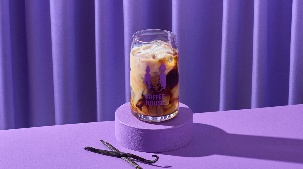 Jääkahvi maidolla lasissa violetin korokkeen päällä. Edessä kaksi vaniljatankoa, taustalla violetti verho.