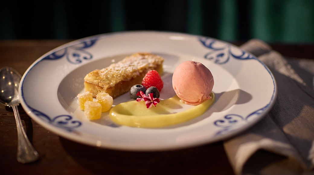 Dessert på en vit tallrik: kaka, jordgubbe, blåbär, rosa glasskula och ljusgul sås.