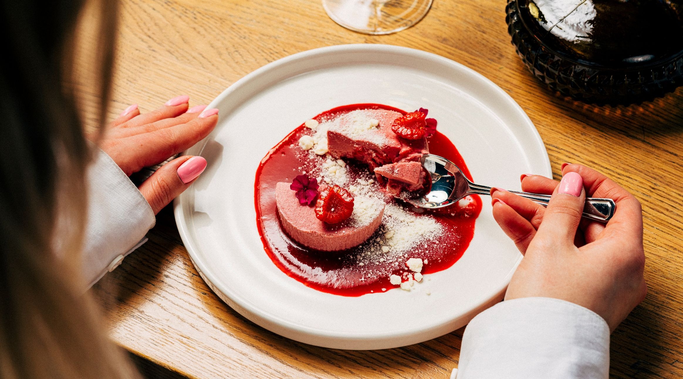 Person äter en rosa efterrätt serverad på vit tallrik med röd sås, garnerad med hallon och florsocker.