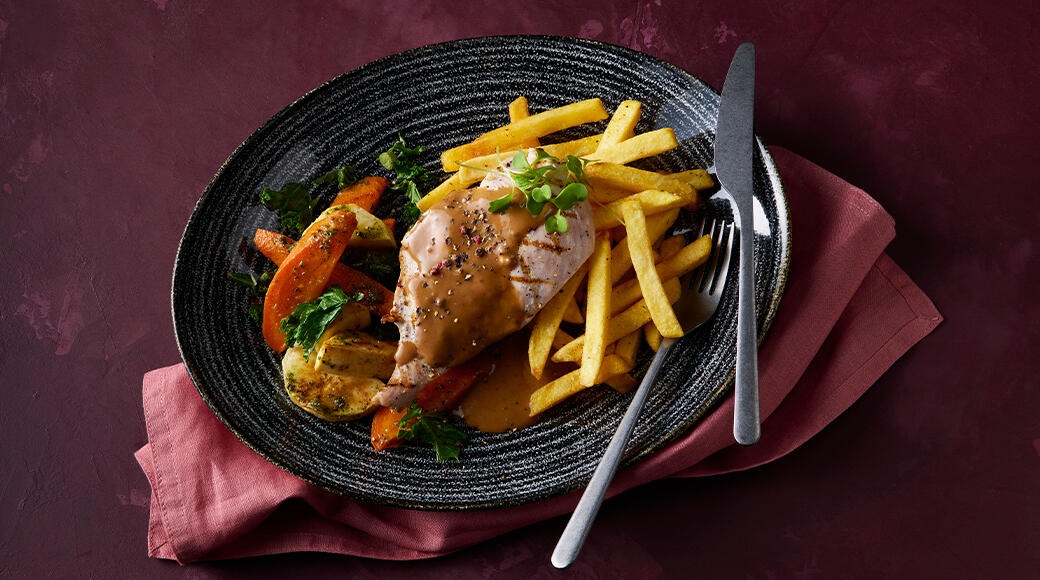 Dark plate with grilled chicken in pepper sauce, french fries, and roasted carrots with herbs. A fork and knife rest on the plate.