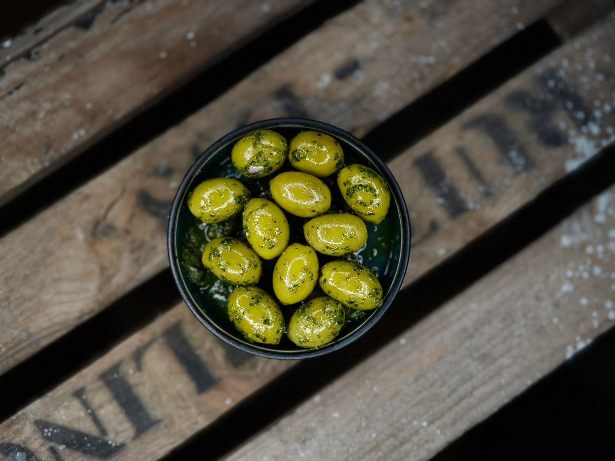 Small bowl of green olives in herb-infused oil on a wooden surface.
