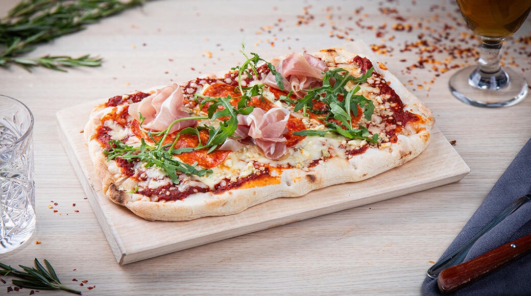 Pizza lautasella, koristeltu kinkkusuikaleilla ja rucolalla, vaalean puupöydän päällä.