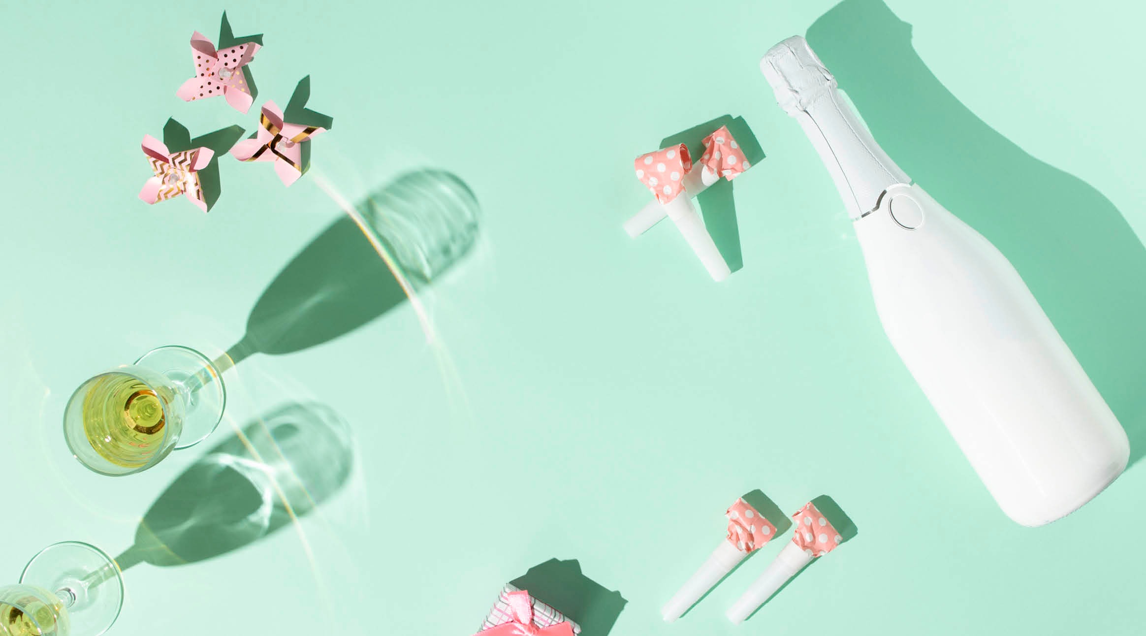Champagne bottle, two glasses of sparkling wine, and festive decorations on light green background. Paper flowers and party straws with pink bows.