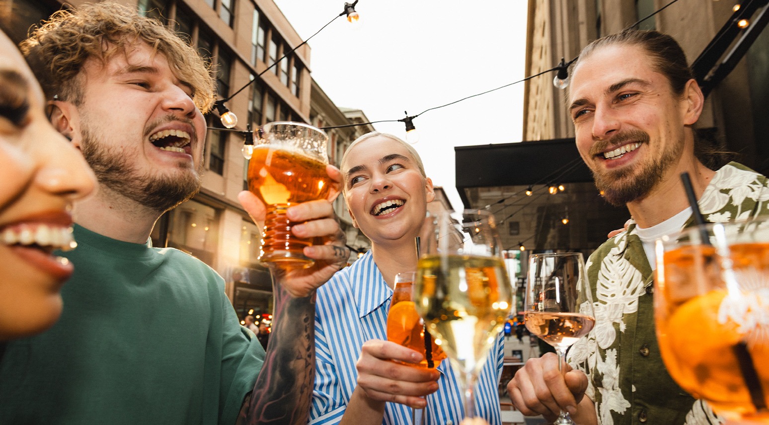Vänner skålar med drinkar på en uteservering med ljusslingor och stadsmiljö i bakgrunden.