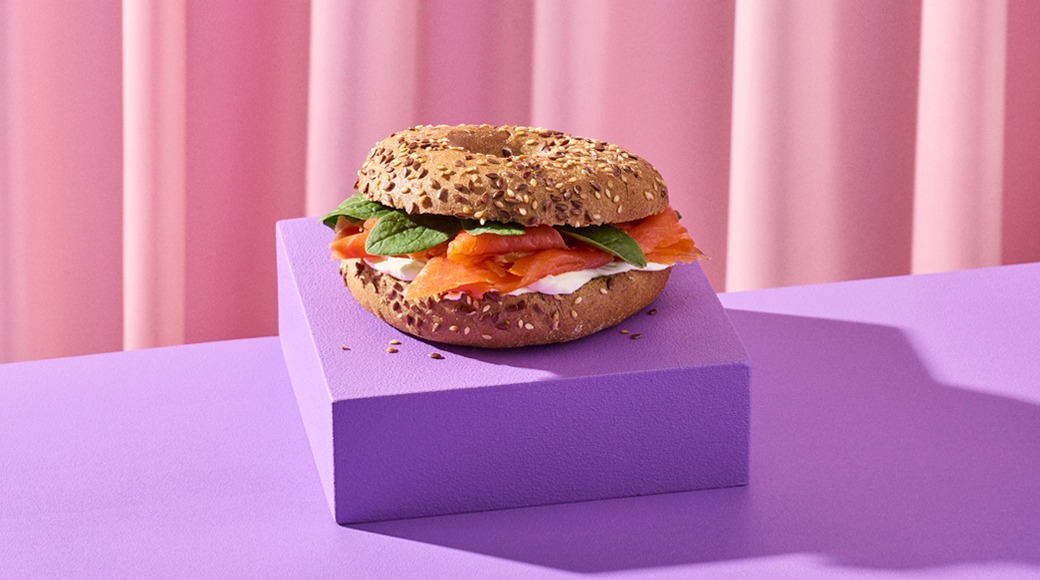 Siemennein kuorrutettu bagel, jossa on graavilohta, tuorejuustoa ja pinaatinlehtiä, violetilla korokkeella vaaleanpunaista verhoa vasten.