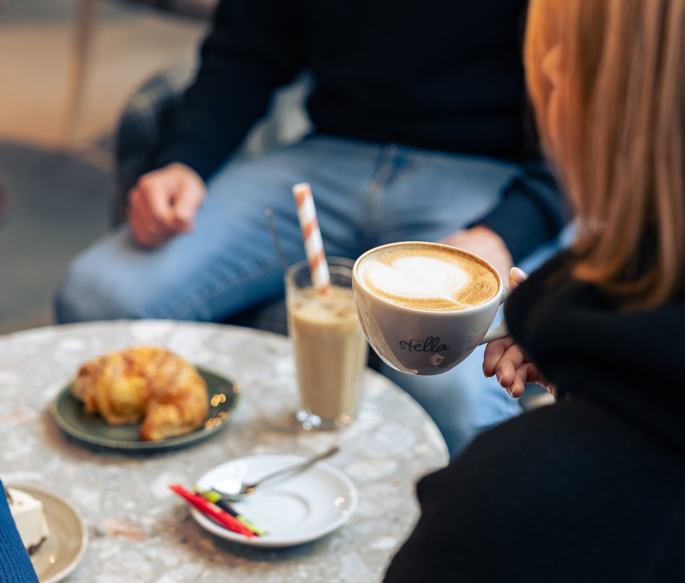 Henkilö pitää käsissään cappuccinokuppia, pöydällä voisarvia ja juomia.