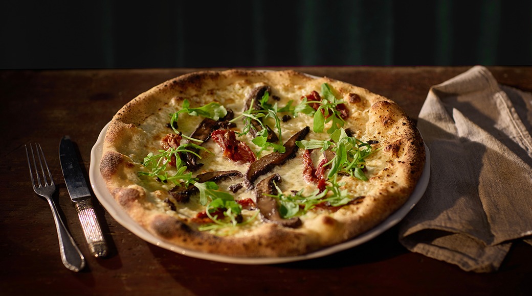 Käsitööpitsa seente, tomatite ja värske rohelisega puulaual, kõrval söögiriistad ja salvrätik.