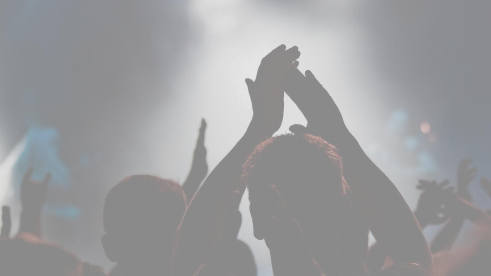 Concert audience clapping and raising their hands. Silhouettes stand out in the dim space, with hazy stage lights in the background.