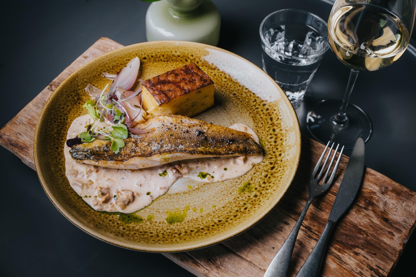 A dish of seared fish and grilled potato in cream sauce. The plate sits on a wooden board next to wine glasses.