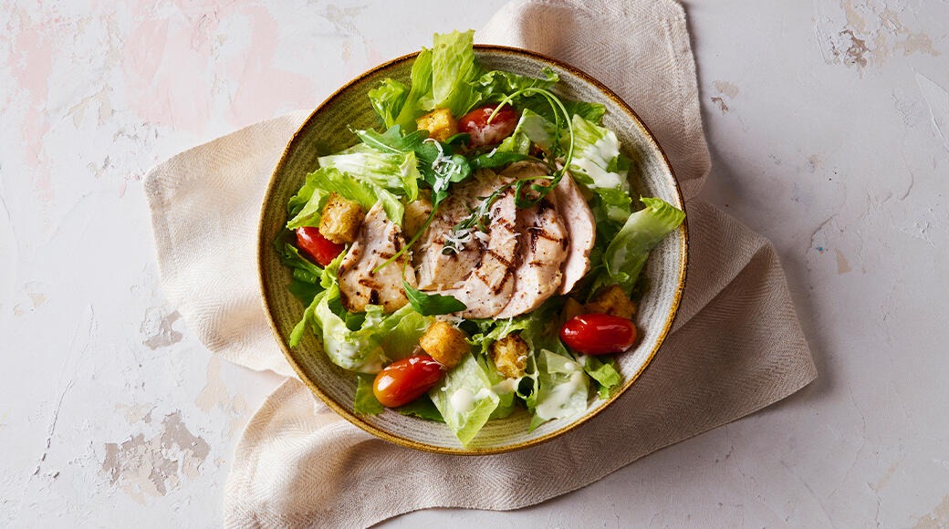 Caesar salad in a bowl with grilled chicken, lettuce, croutons, cherry tomatoes and dressing. The bowl rests on a light cloth.