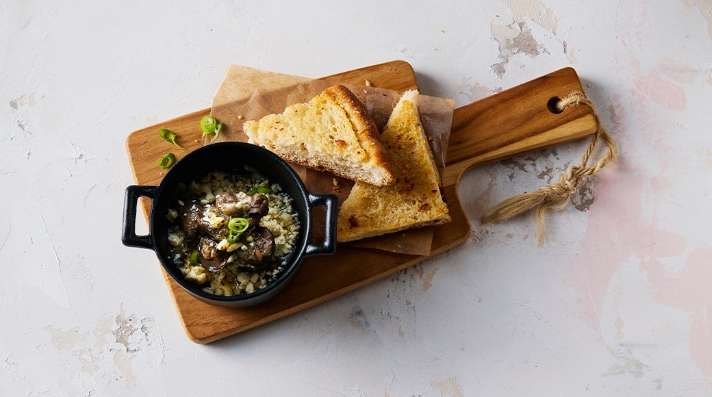 Small black cast-iron dish of garlic snails topped with spring onion. Toasted bread slices sit beside it on a wooden serving board.