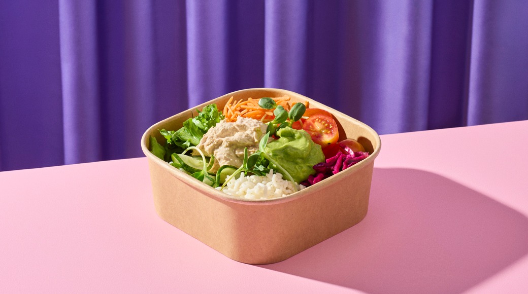 Kartonkirasiassa on salaattiannos, jossa on riisiä, guacamolea, hummusta, porkkanaraastetta, tomaattia, punakaalia ja versoja. Rasia on vaaleanpunaisella pöydällä violetin verhon edessä.