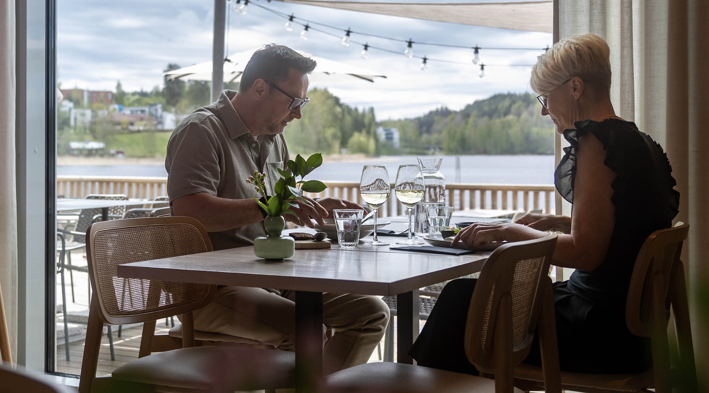 Ravintolan pöytä lasiseinän edessä, näkymä järvelle ja vihreälle maisemalle. Taustalla avoin patioterassi lasioven kautta.