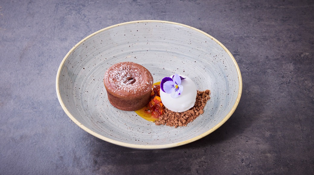 Vackert presenterad dessert på en ljus tallrik: chokladkaka, vit glass dekorerad med lila blomma och chokladsmulor.