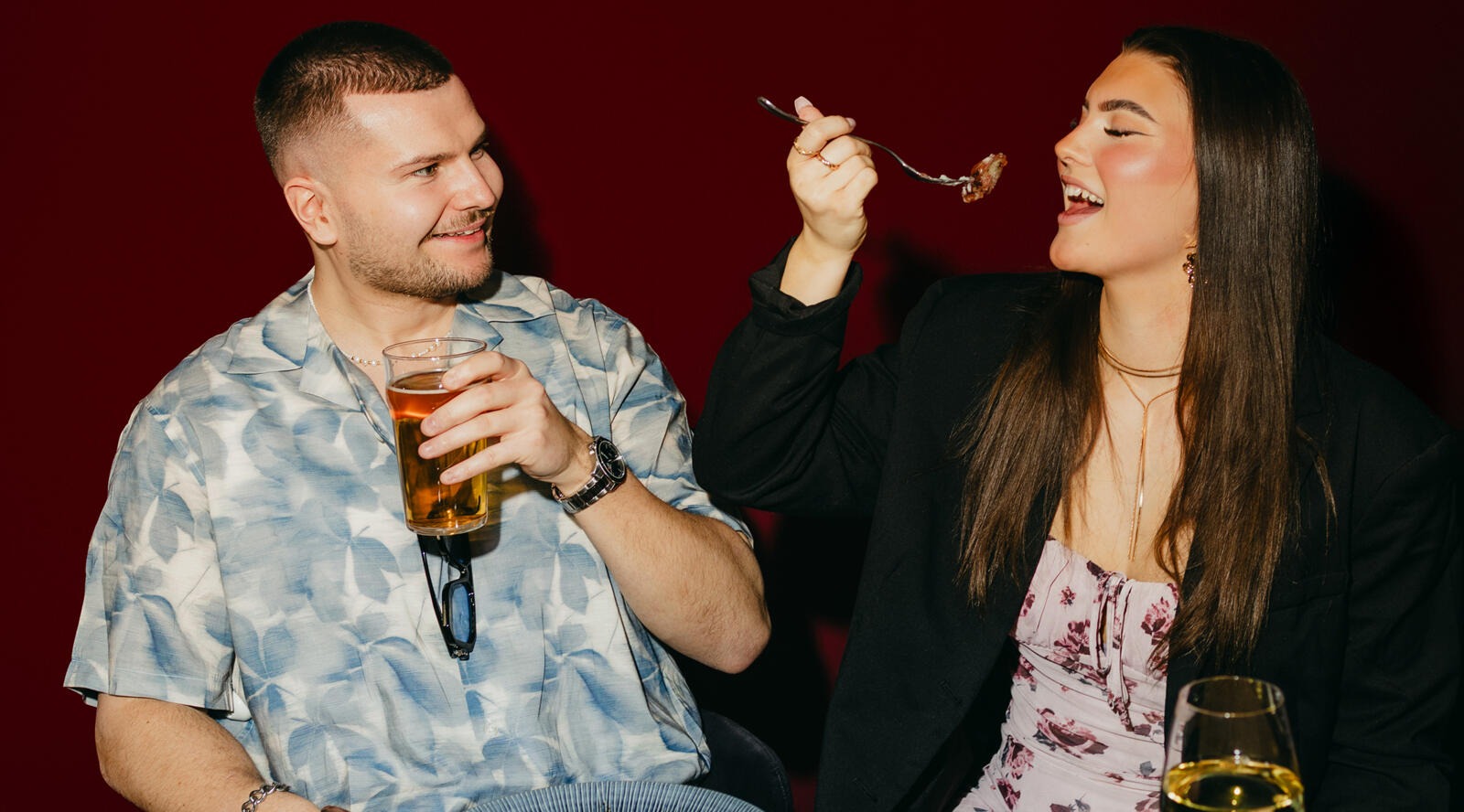 Två personer som njuter av mat och dryck i ett rum med röd bakgrund. En håller i ett ölglas, den andra äter med sked.